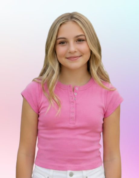A young blonde girl wearing a pink short-sleeve henley t-shirt and white pants, standing against a pastel gradient background.