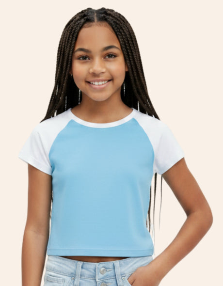 A young girl with long black braids smiling while wearing a light blue and white raglan crop top and light-wash jeans, set against a solid pale beige background.